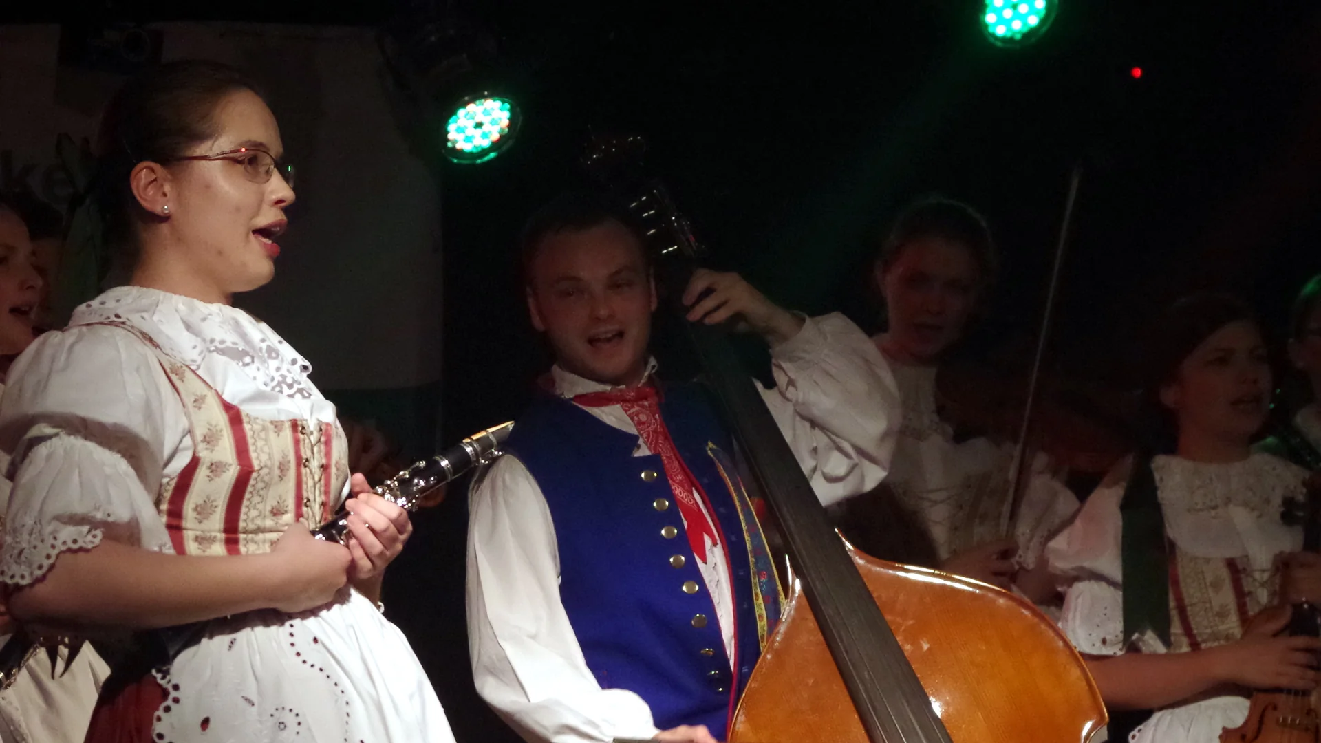 A Czech folk band in traditional costume performing on stage in the evening — clarinet, singer and double bass, representative of a Czech folklore evening